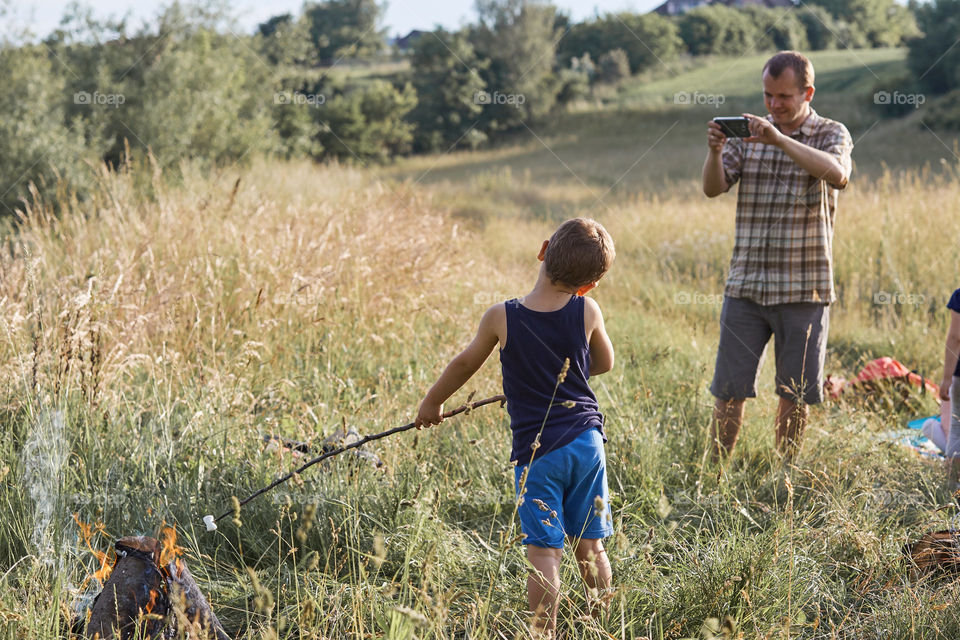 Man taking a photo his son roasting marshmallow over a campfire. Family spending time together on a meadow, close to nature. Candid people, real moments, authentic situations