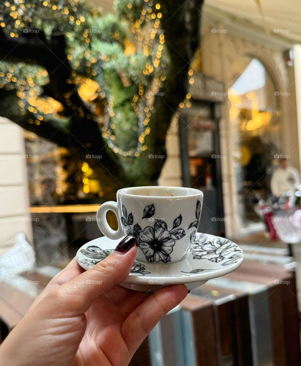 Woman’s hand with dark nails holding small white and black coffee cup. Bright light, tree and the street in the background 
