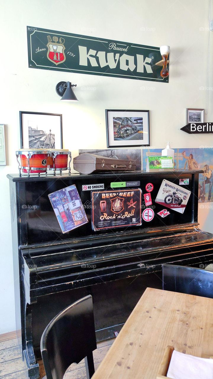 Cafe interior. An old black piano covered in stickers. On the lid of the piano stands a Bongo and a violin case. Fotos hanging on the wall. Retro style