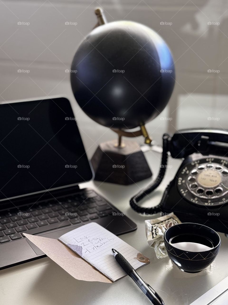 Glass desktop with iPad, globe, vintage desk telephone, black tea cup with small spill and crumpled napkin, small note booklet and black ball point pen