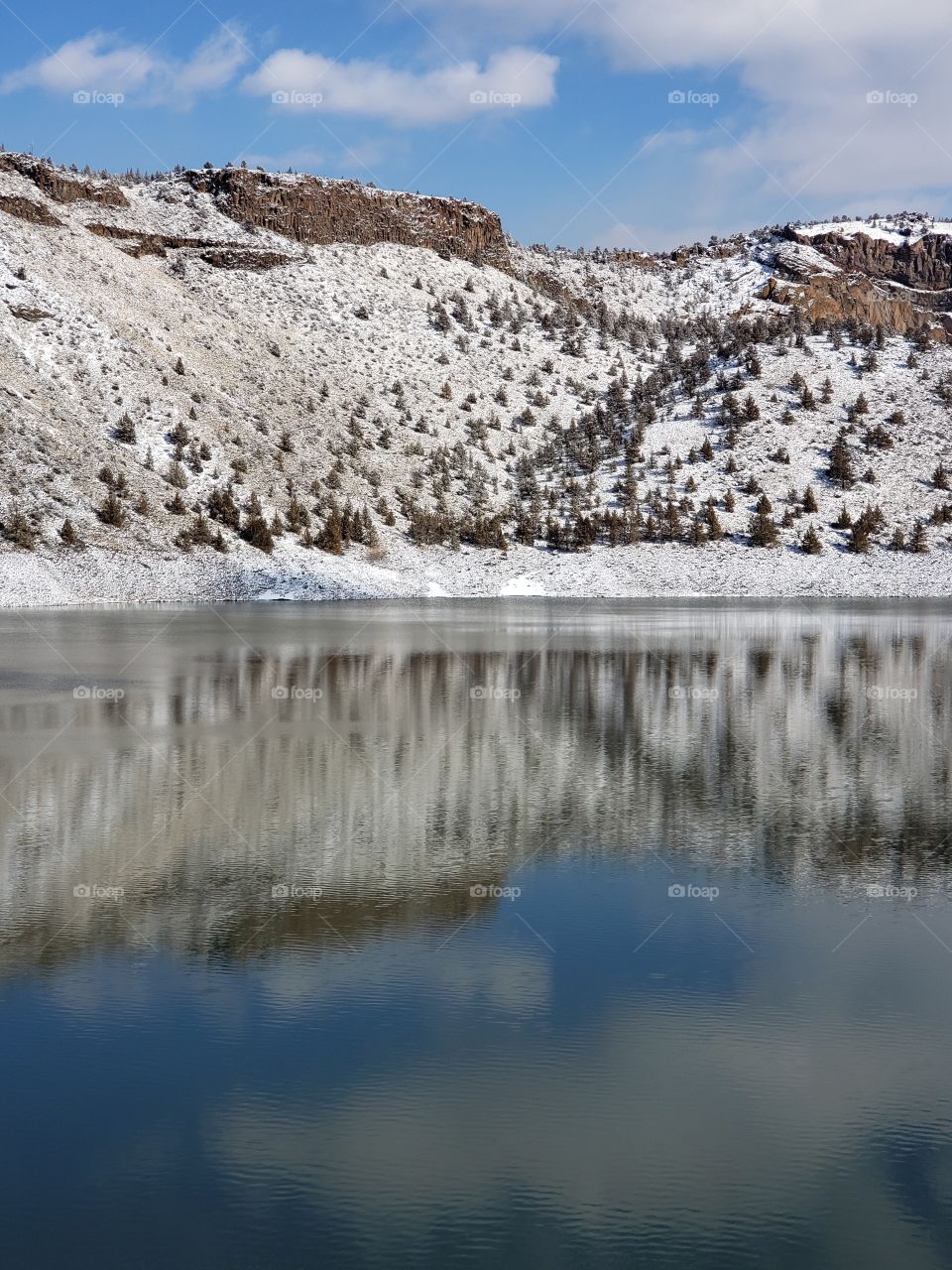 Snow covered hills, trees, clouds, and a bright blue sky reflect in the partially iced over Prineville Reservoir on a sunny winter day in Central Oregon. 