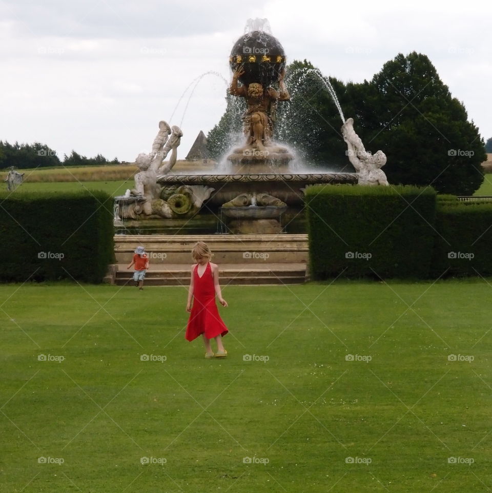 A young girl in a beautiful red dress walks along manicured lawns with a magnificent fountain in the background all part of the landscaping at Howard’s Castle outside of York in England on a summer day. 