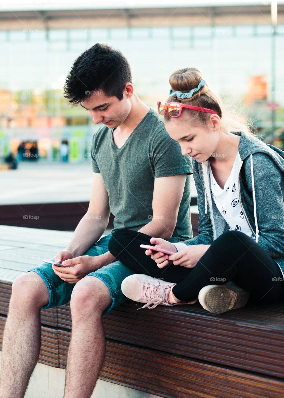 Couple of friends, teenage girl and boy,  having fun together, using smartphones,  sitting in center of town, spending time together. Real people, authentic situations