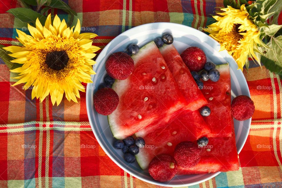 Watermelon with red berries on a plate and two sunflowers on a picnic blanket.