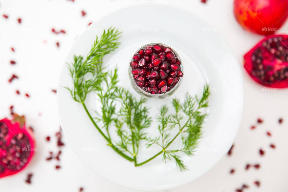 Delicious fresh and clean image of pomegranate seeds, fruit and herbs on a white plate and scattered seeds on a white background. Delicious and healthy.