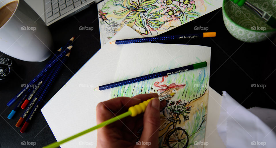 Closeup of hand holding Faber-Castell Aquarelle Colored watercolor pencils sketching girl on bicycle in countryside with sketchbook on table with laptop and coffee conceptual work life balance, creativity, hobby artist lifestyle photography 