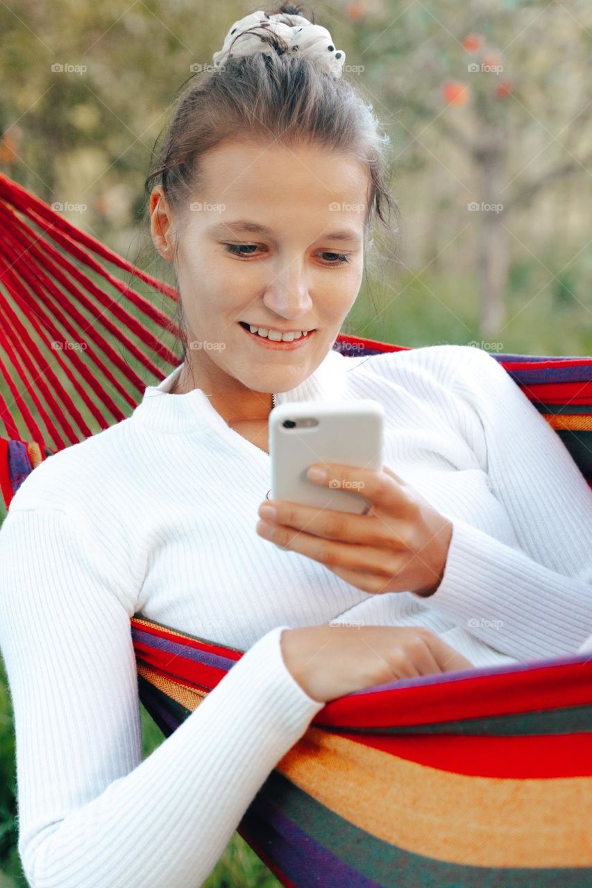 Millennial woman using phone texting chatting in social media networking while resting in hammock 