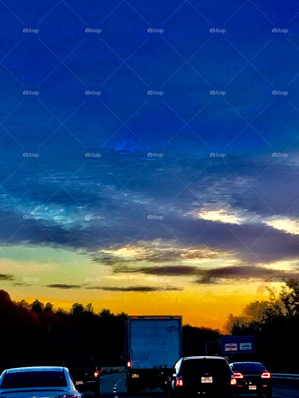 Highway view in traffic jam of the sunrise/sunset. The low sun lights up the clouds in beautiful shades of color. Highway is lined with woodlands and vehicles have headlights & break lights on... we're not moving.