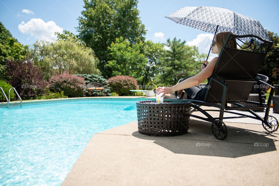 From behind view of a young millennial woman sitting poolside in a lounge chair and holding an umbrella