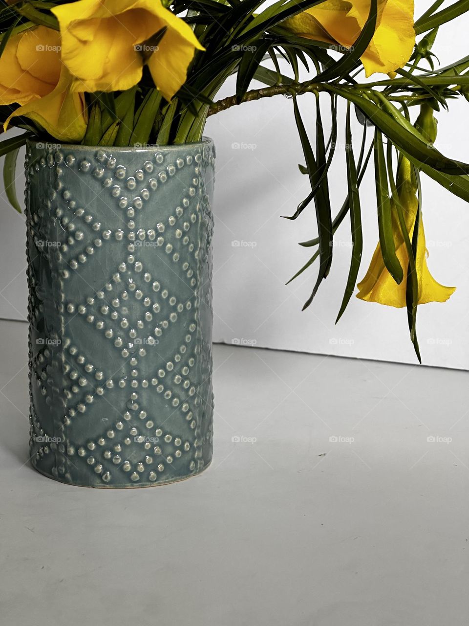 Yellow trumpet shaped flowers with green leaves in a grayish green patterned ceramic vase