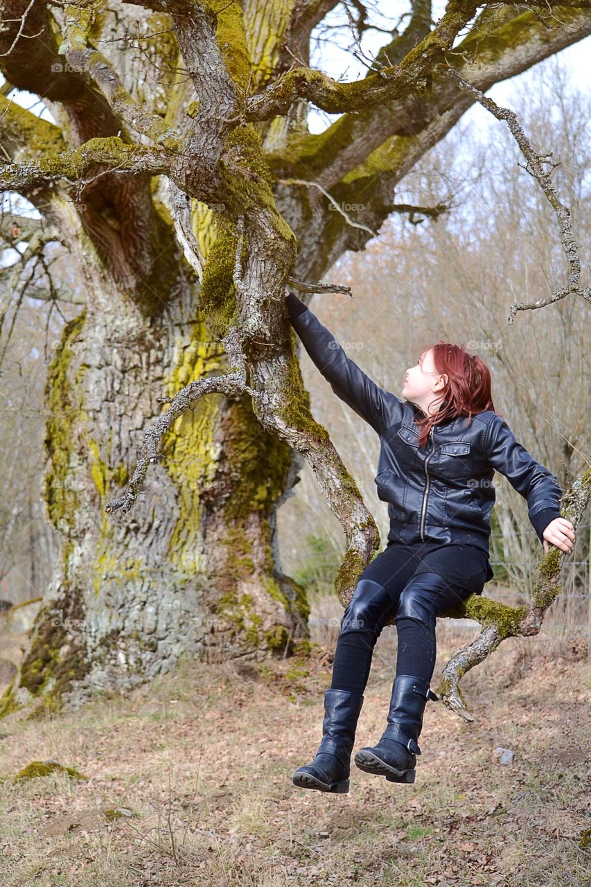 Girl sitting on a tree branch in leather jacket and black pants enjoying the life