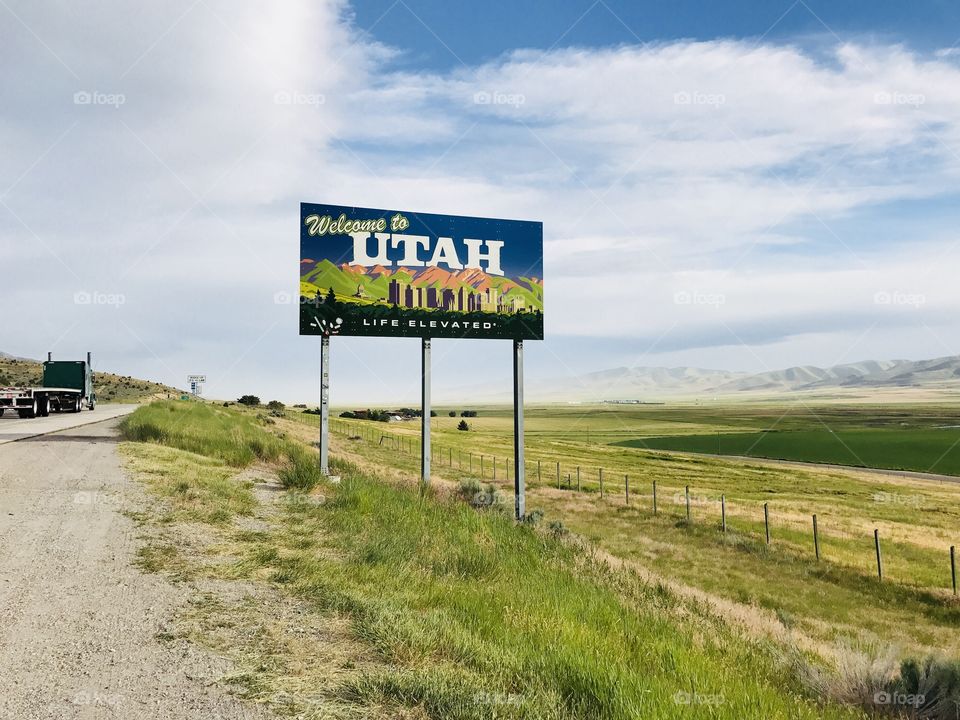 The welcome to Utah sign is against a bright cloudy and blue summer sky with the mountains in the background. 