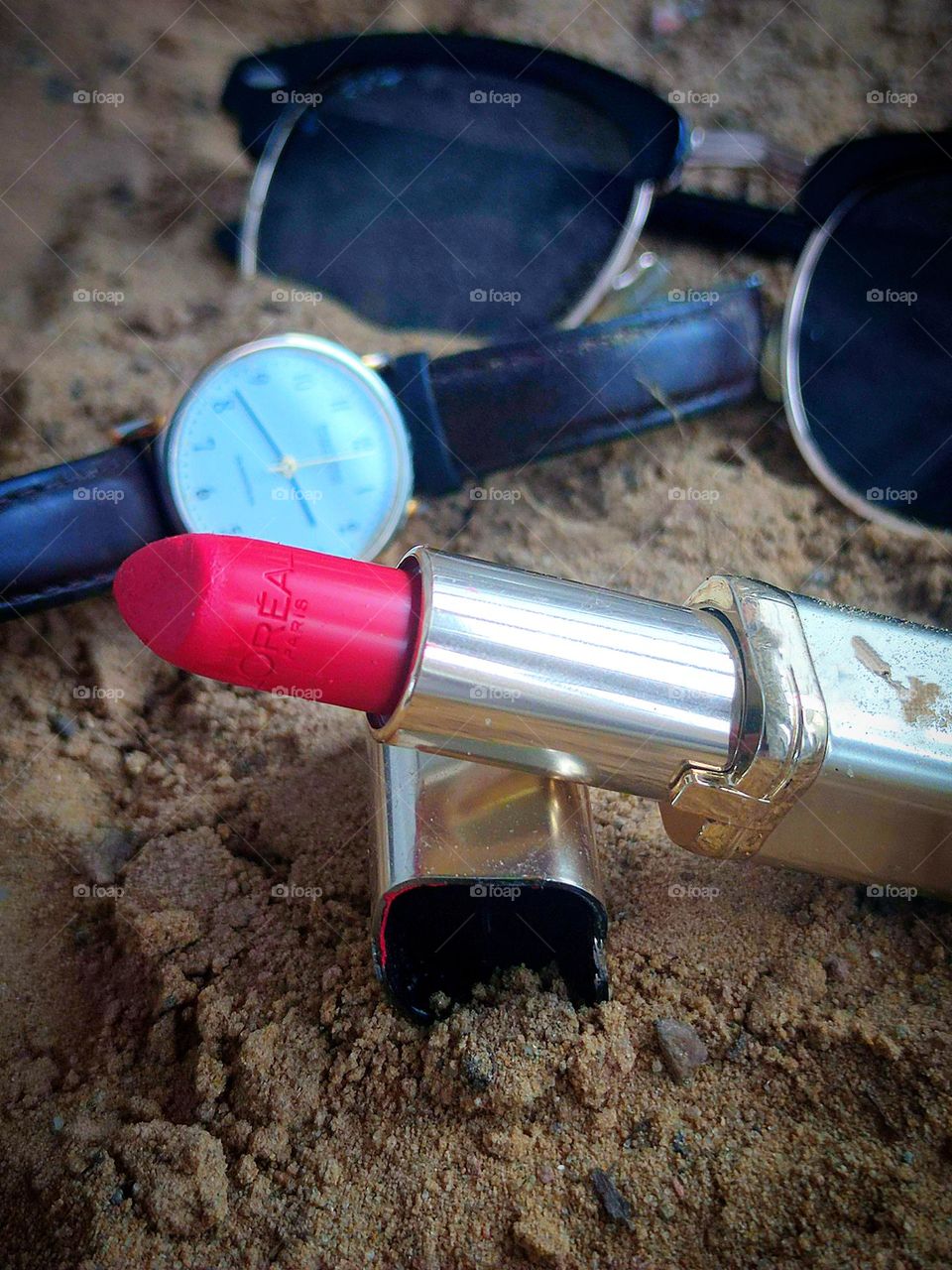 There is a lipstick cap on the sand with a tube of open red lipstick leaning against it.  In the background, a wristwatch and sunglasses lie on the sand.