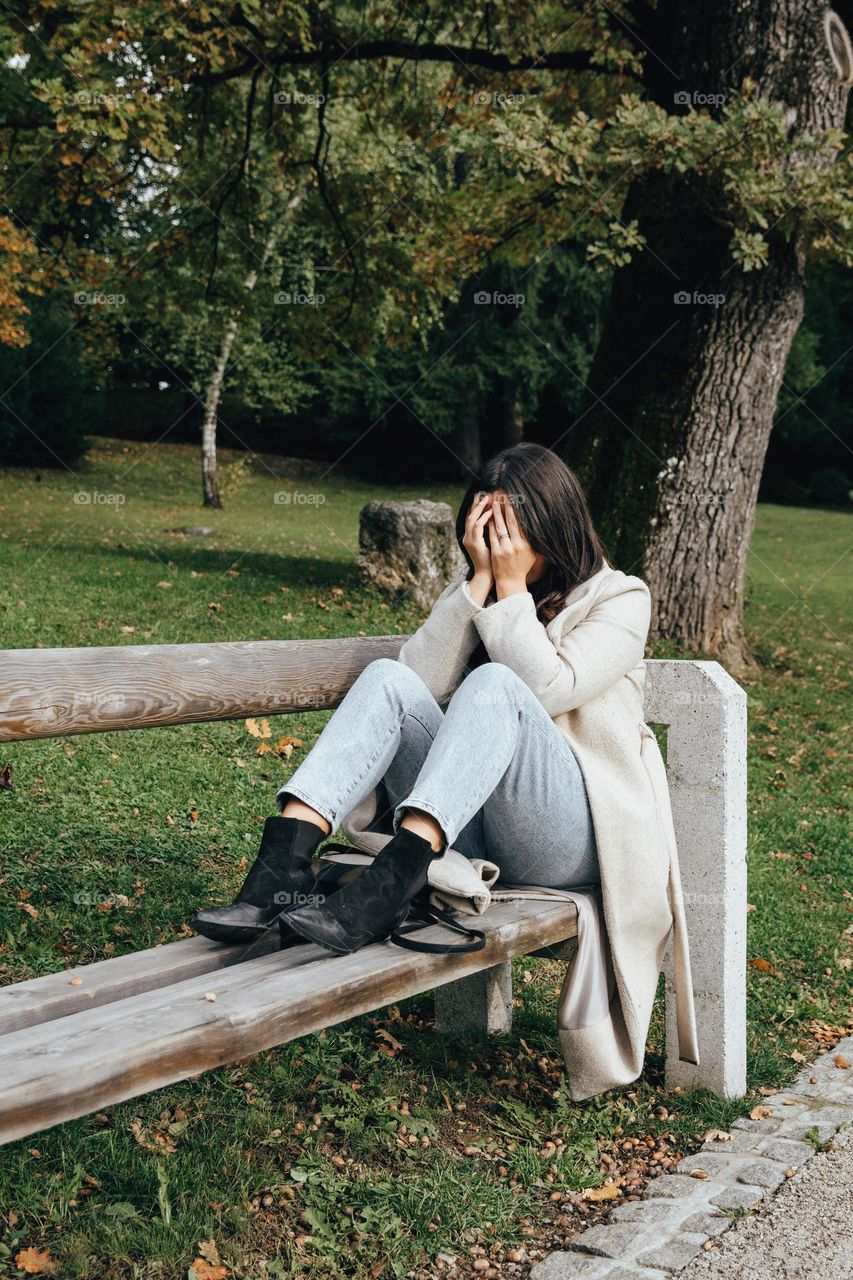 Sad woman sitting on bench in park, holding hands over her face