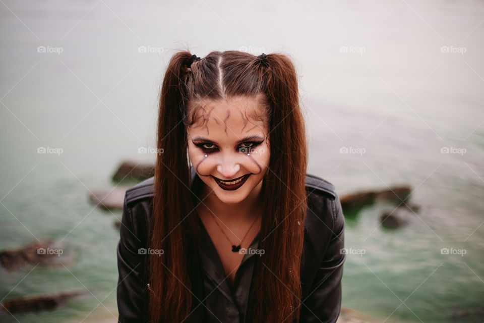 Cheerful scary woman with Halloween makeup with pale face wears costume for carnival party, posing against the backdrop of water, having fun at the holiday. A creative idea for a masquerade.	