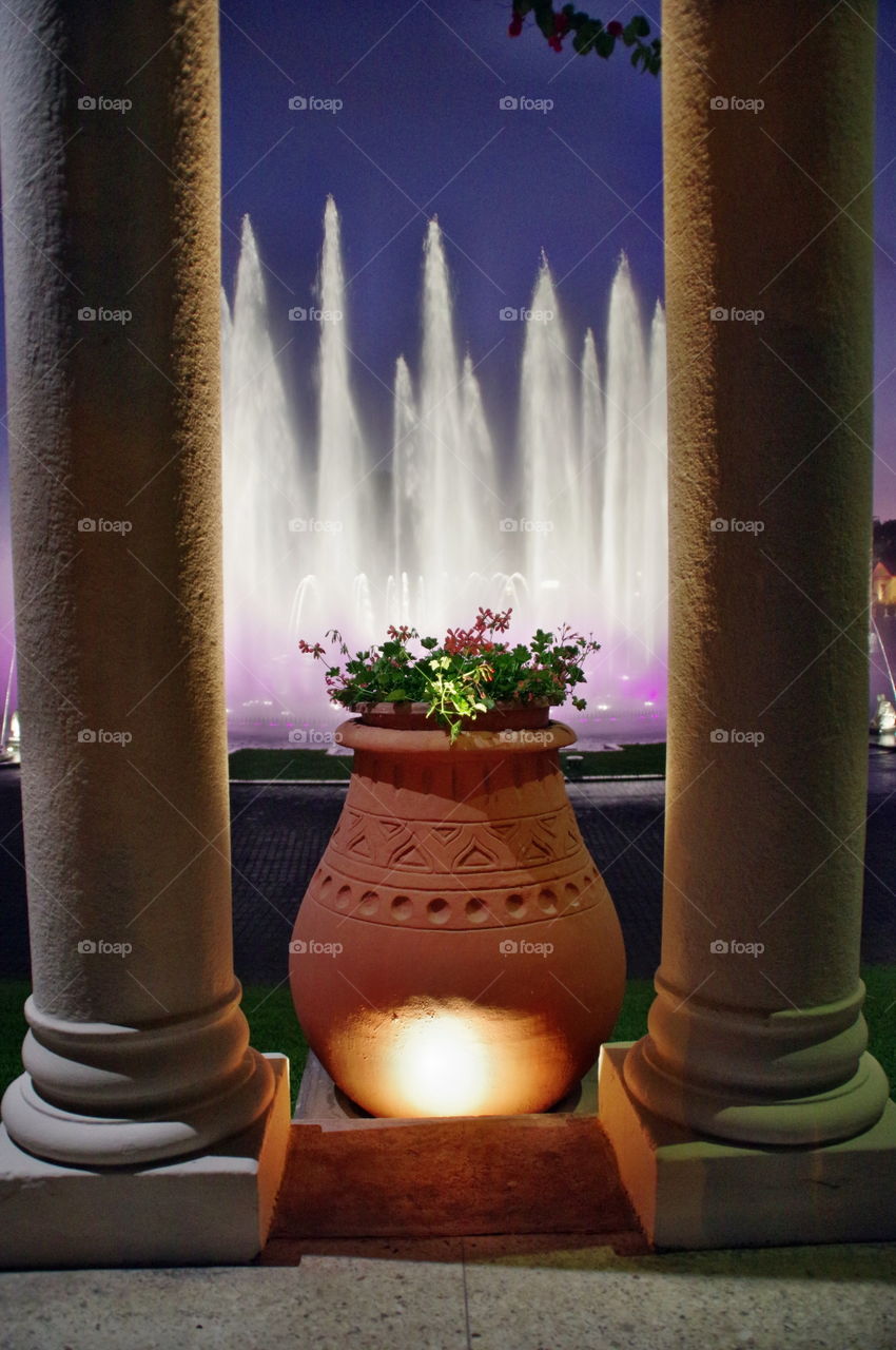 Big clay jug with flowers surrounded by two pillars against a colorful water fountain