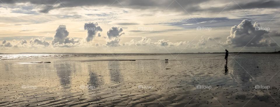 A man walks a dog on a wet beach, reflecting the clouds in the sky - West Wittering Beach, UK