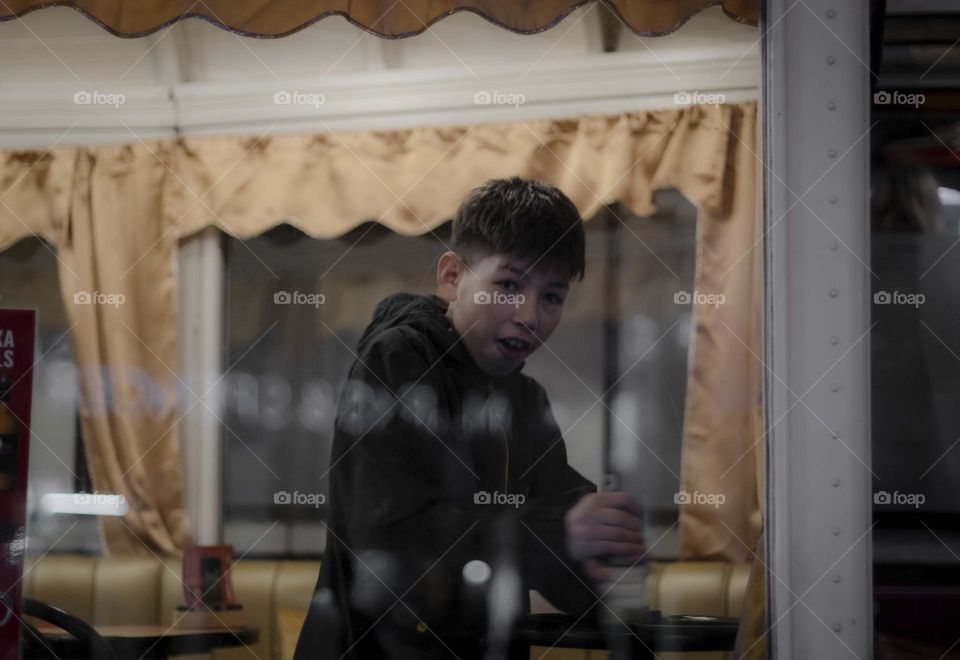 Portrait of a handsome caucasian brunette teenager boy in a dark blue jacket with a short haircut standing outside the window in the restaurant carriage and spinning a wooden wheel with an emotion of surprise looking at the camera, side view close-up