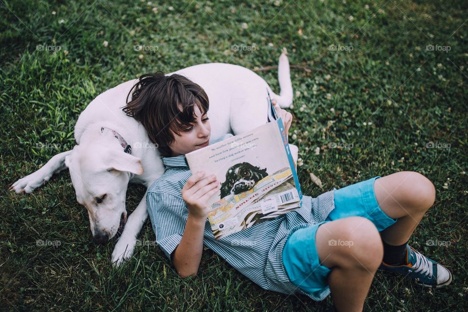 Young boy laying down on his dog reading a book