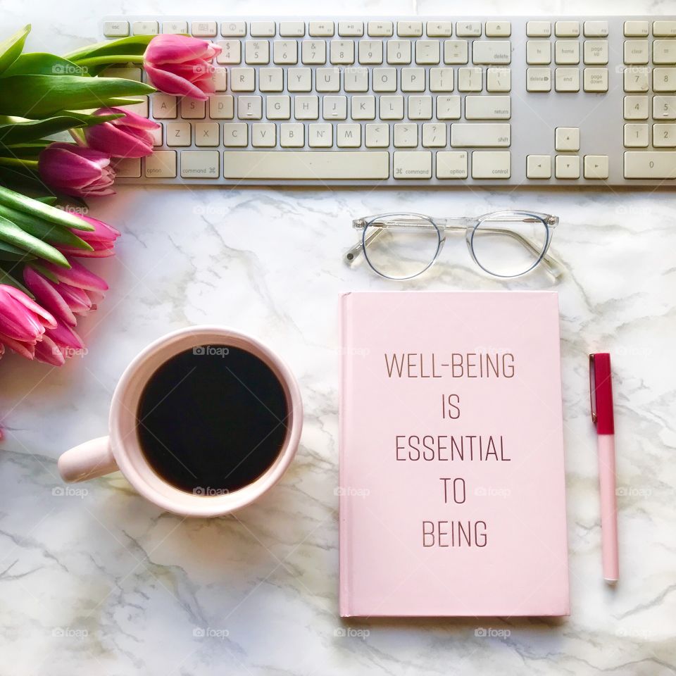 Well being is essential to being! Take care of yourself! A pink book on a marble table with a pink coffee cup and white keyboard!
