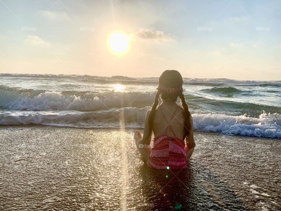 Little girl sitting in meditation position in the surf during sunset 