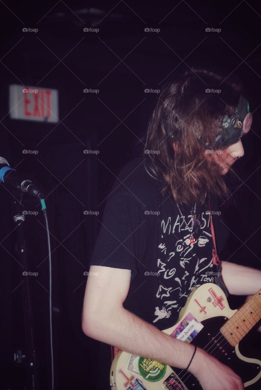 Man with long wavy hair wearing a bandana plays guitar and looks down while he strums and stands in front of a microphone. 