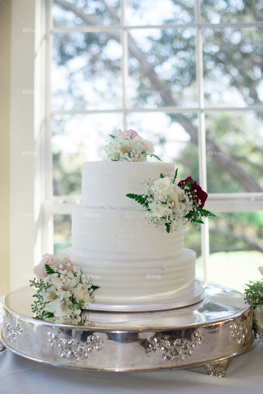 White three tier cake with pink and red roses near window on silver cake tray