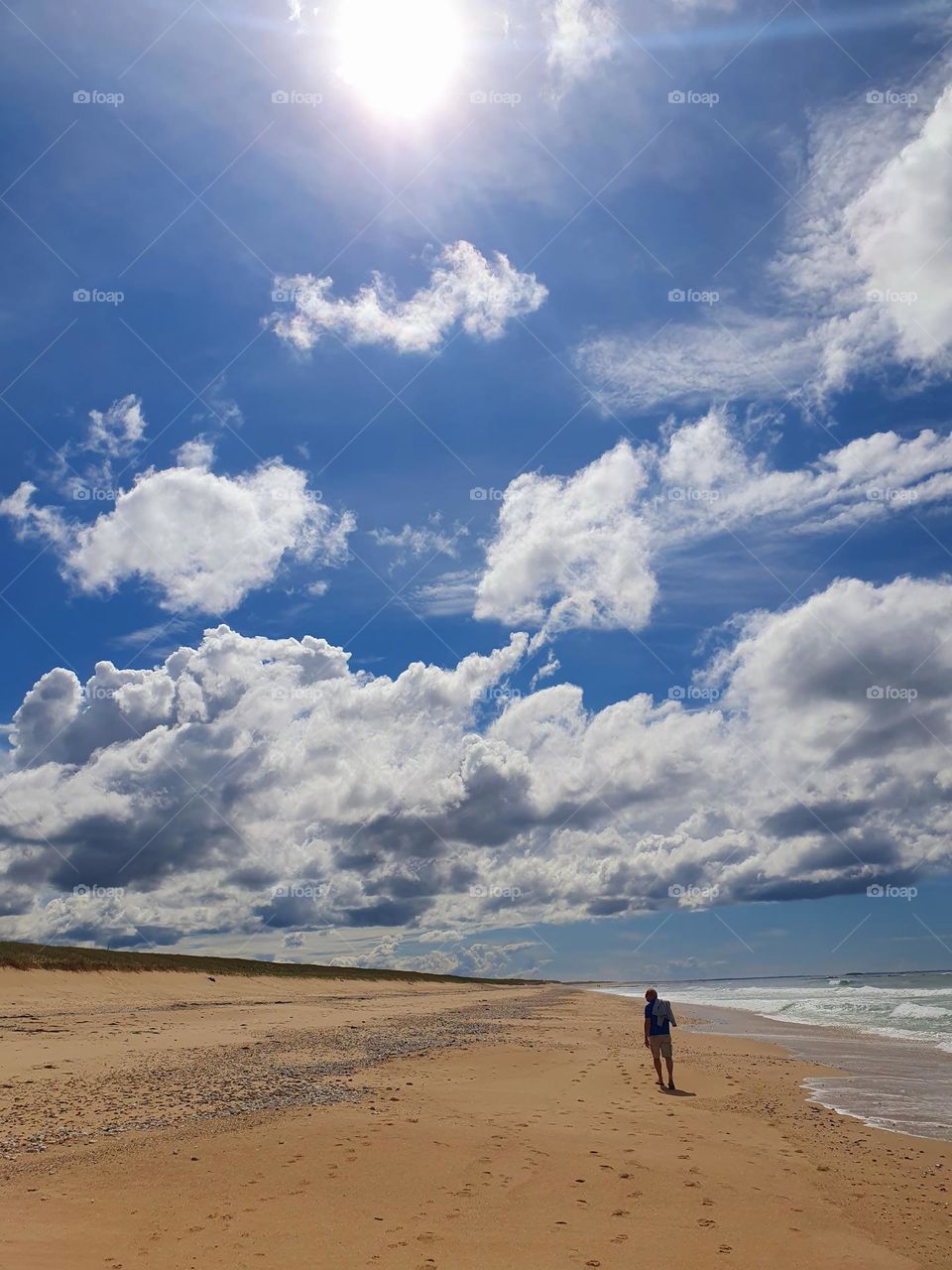 A father walking on the seaside of Mât Fenoux dunes under a bright blue sky with white clouds and white shining sun