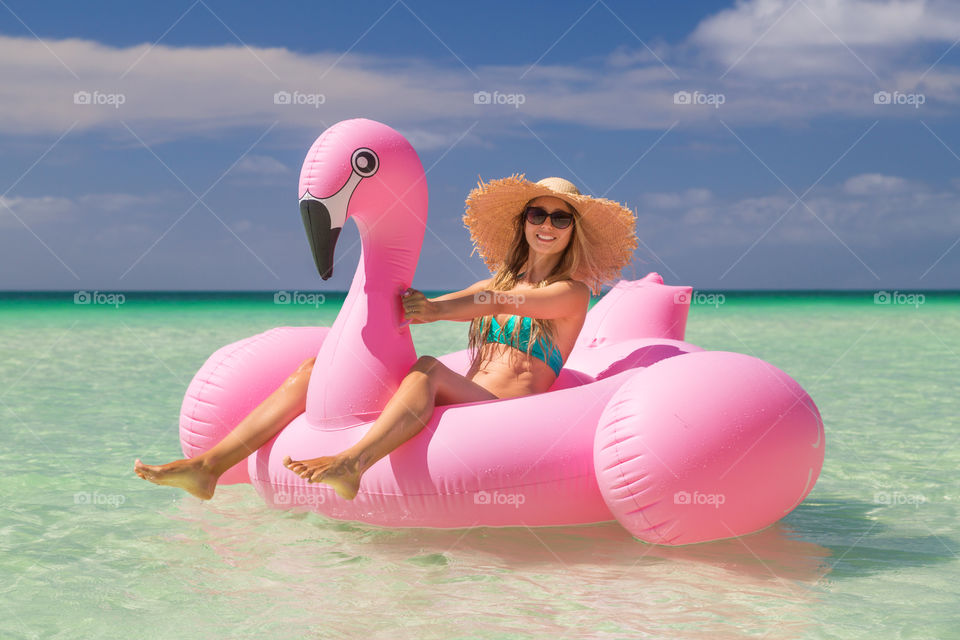 Happy young woman with long blonde hair in bikini, sunglasses and straw hat having fun ob the sea with inflatable flamingo 