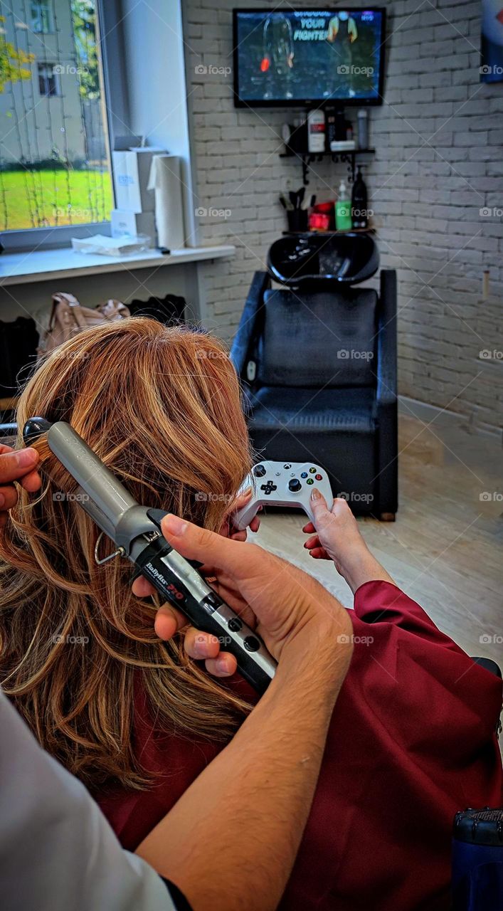 Video games. Beauty saloon. A woman sits in a chair and plays a video game. Hands of a hairdresser doing a woman's hair