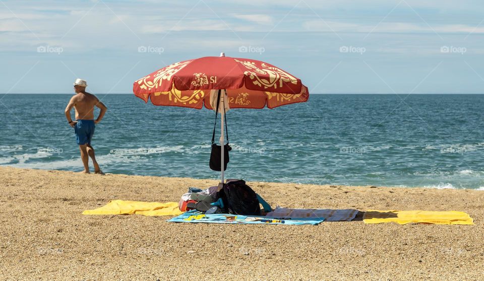 A beach parasol shelters a pile of bags, clothes, towels and flip flops on the beach, as a man considers a dip in the sea