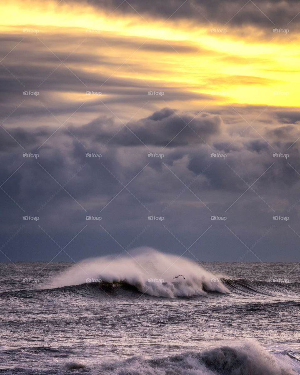 Dark ominous storm clouds taking over warm golden hour light, as white spray comes off large breaking waves.