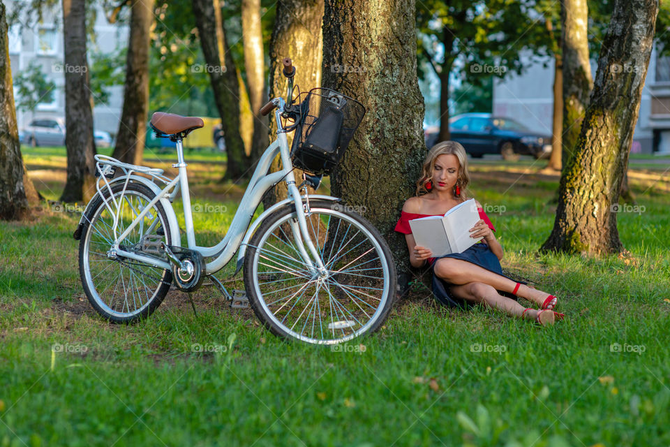 a young blonde woman with a white bike sitting in a park near a tree and reading a book