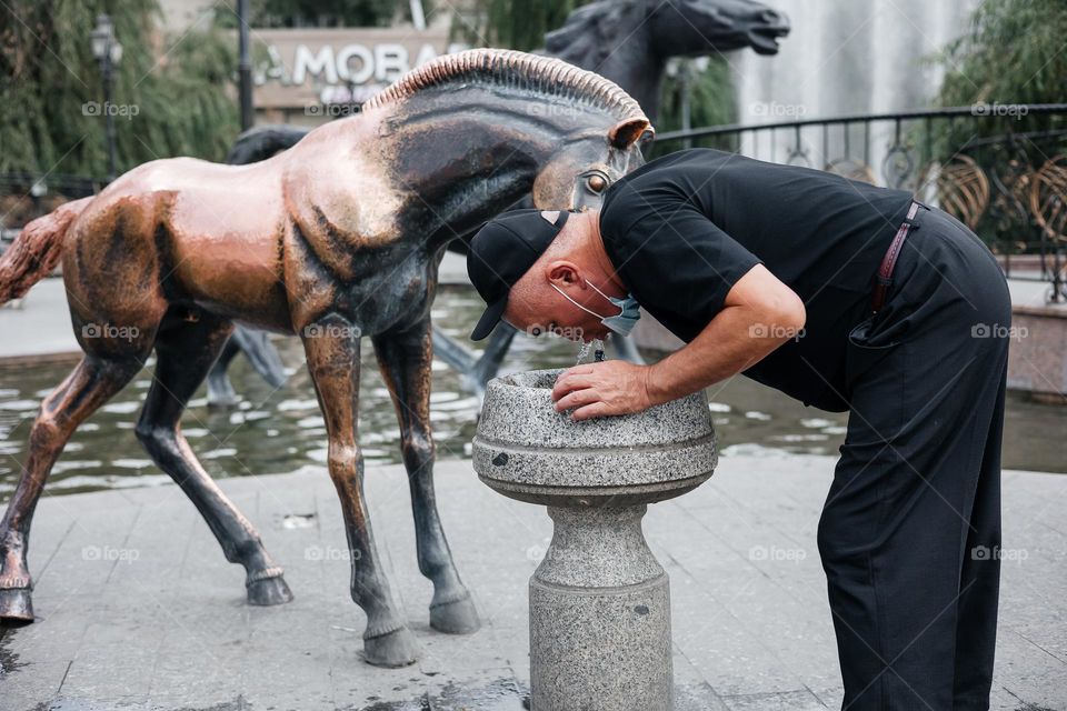 Asian man in a black wear with his face mask pulled down, drinking water from a fountain.