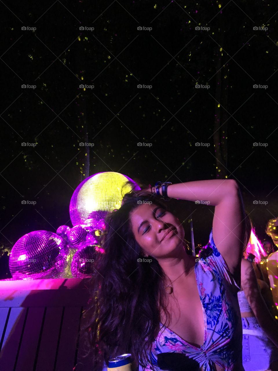 A festival girl poses in front of a disco ball arrangement. It’s the end of the night. The disco balls reflect purple & gold light into the tall dark trees in the background. Her eyes closed, dark hair to the left, a hand resting behind her head.