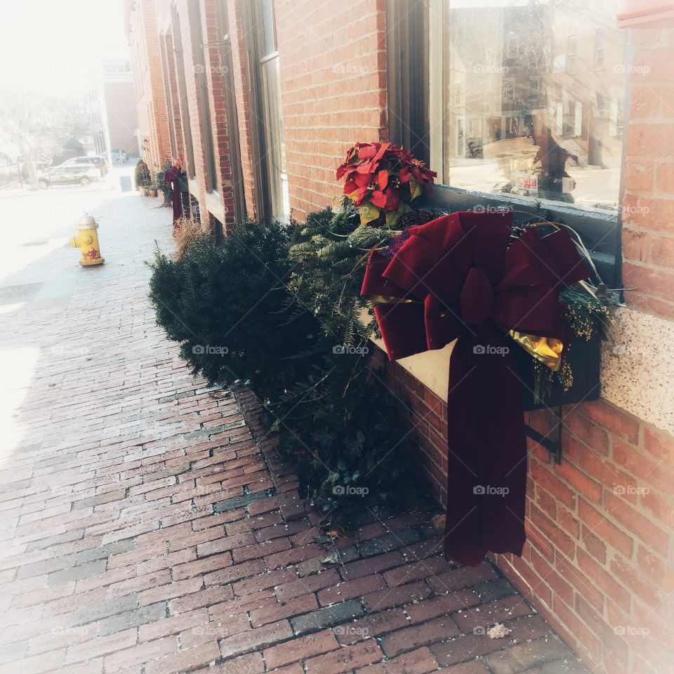 A dark red Christmas bow sits on a window box that has been decorated with poinsettias and evergreen boughs.
