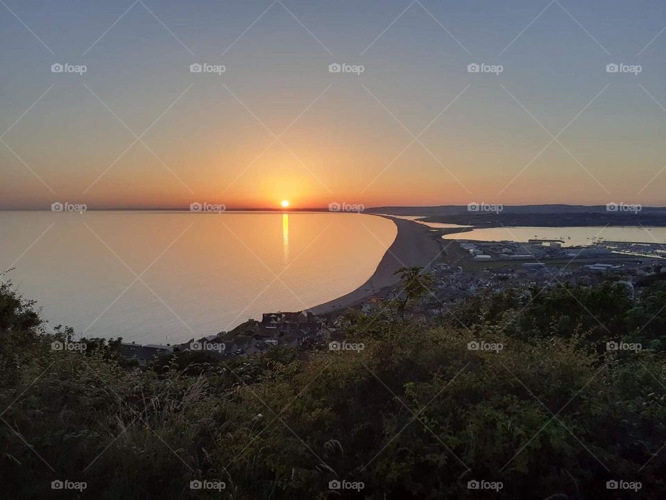 A VIEW FROM PORTLAND IN DORSET LOOKING ACROSS OVER CHESIL BEACH AND THE SUN SETTING IN THE DISTANCE
