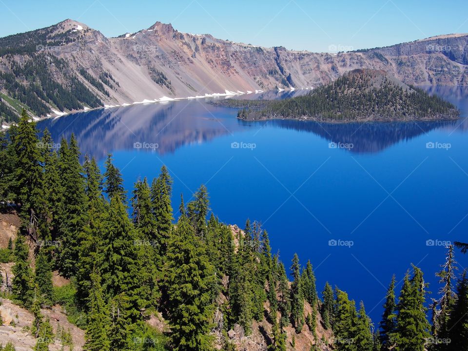 The jagged rim reflecting into the rich blue waters of Crater Lake in Southern Oregon on a beautiful summer morning with perfect clear blue skies.