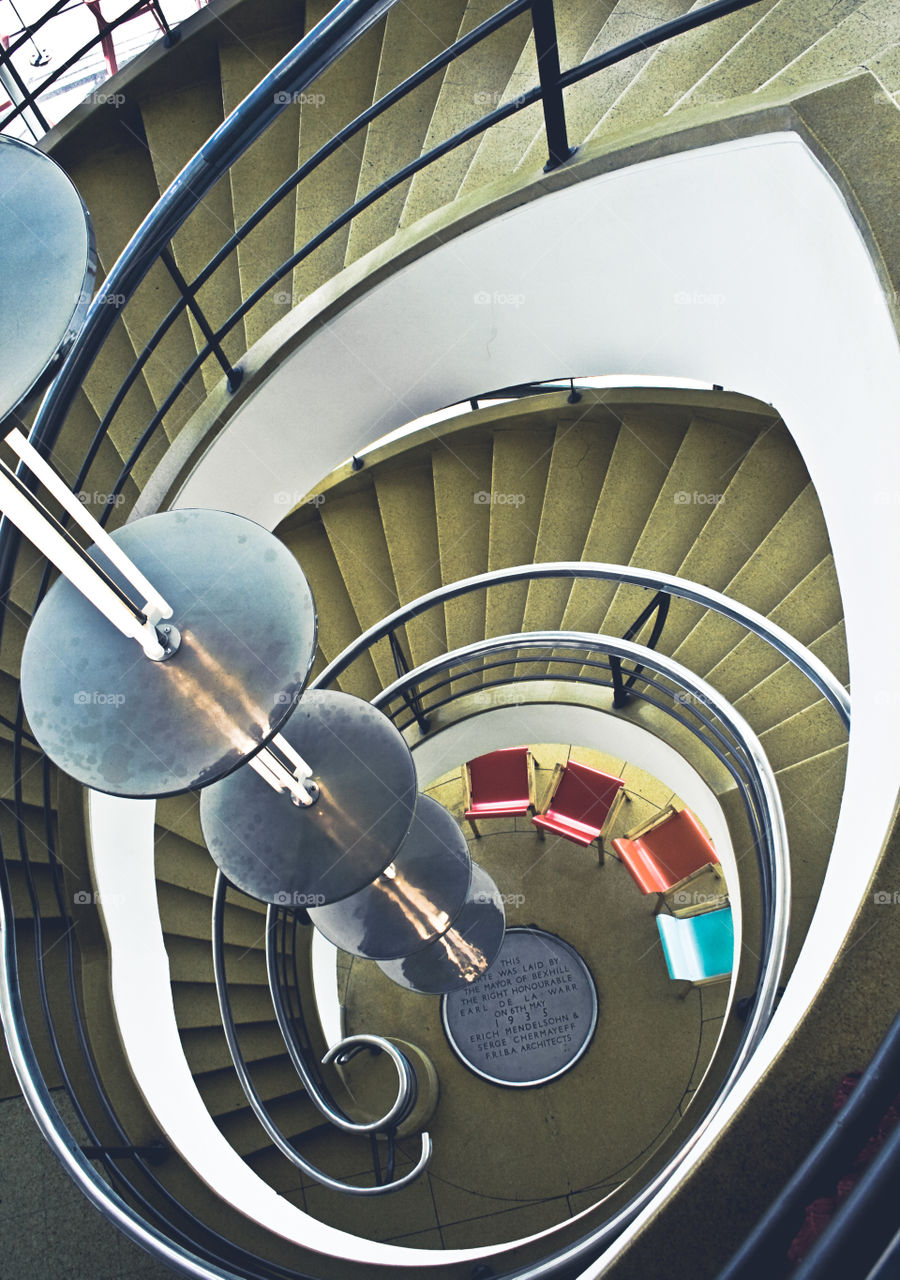 View down a spiral staircase, past a metallic light fixture down to colourful plastic chairs on the ground floor