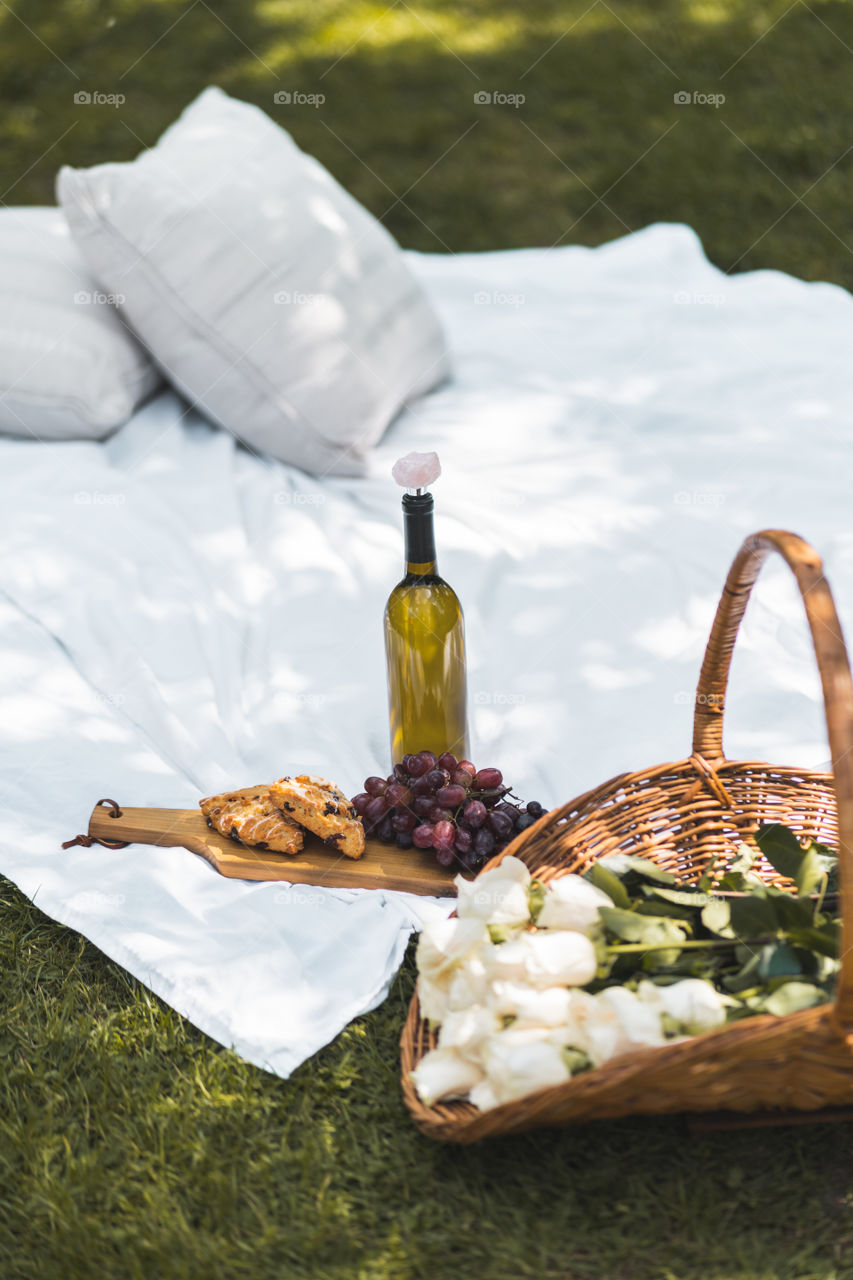 An outdoor picnic setup with flowers, danishes, fruit & wine with pillows and a picnic blanket for summer activities 