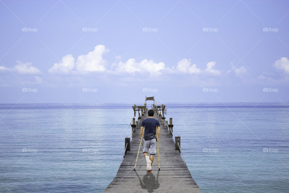 Asian man used wooden crutches walks on Wooden bridge pier boat in the sea and the bright sky at Koh Kood, Trat in Thailand.