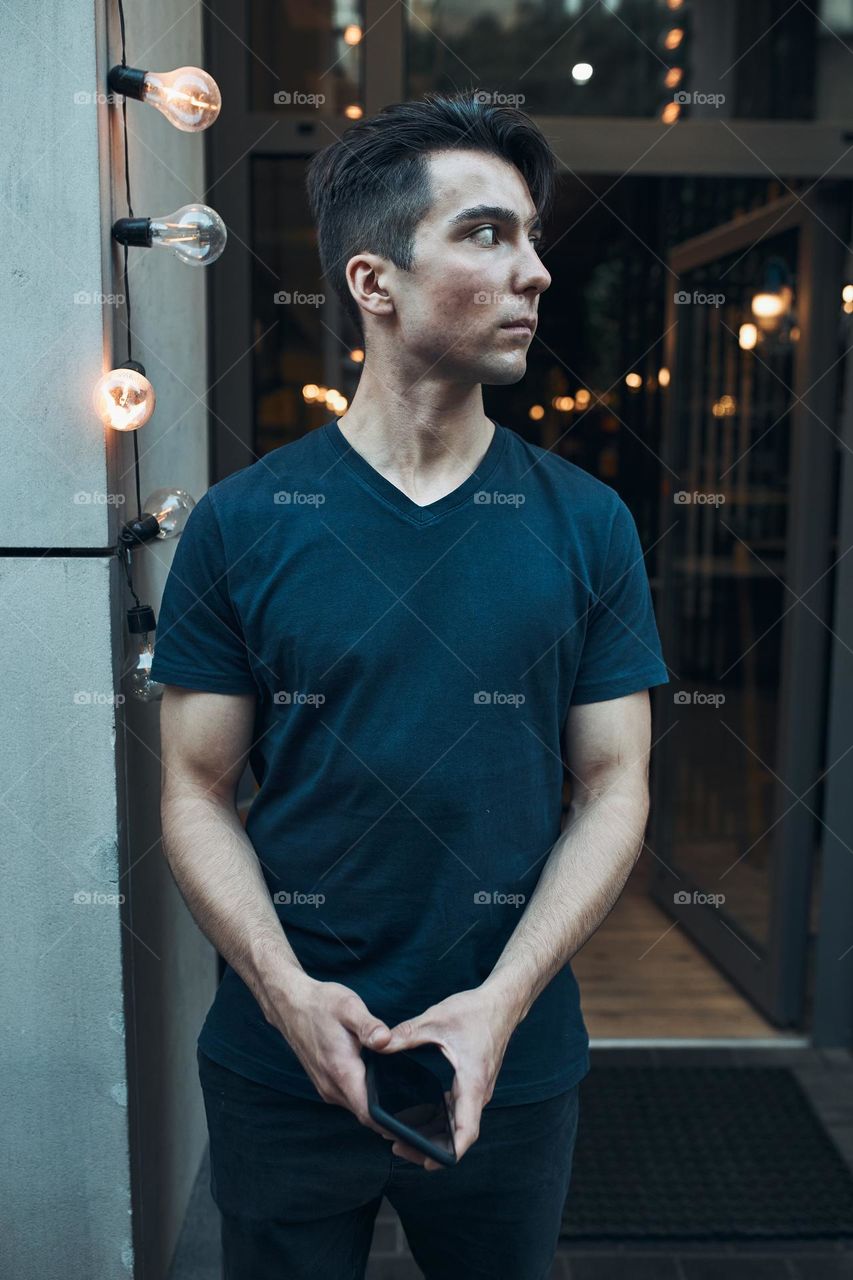 Young man standing in front of restaurant in the city center holding smartphone looking away