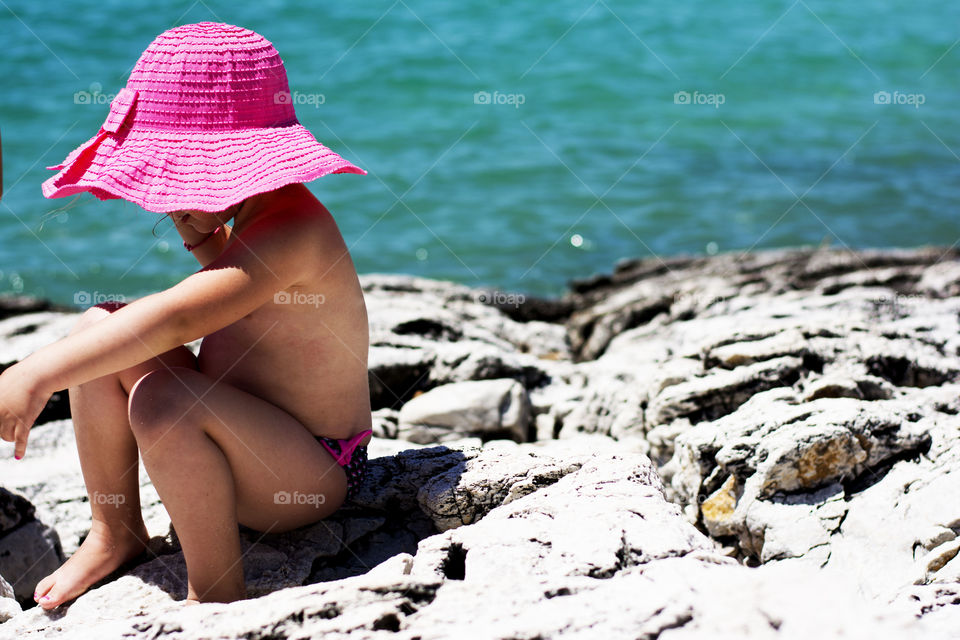 little girl with pink hat. little girl with pink hat sitting on rocks with turquoise sea in the background