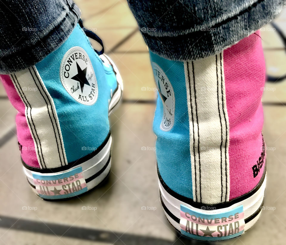 The back (ankles/heels) view of a pair of bright pink, blue & white high-top canvas shoes. Visible Converse All Star logo patches on inner ankles and logo printed on rubber transgender pride flags at bottom of each shoe. Grey tile floor out of focus.