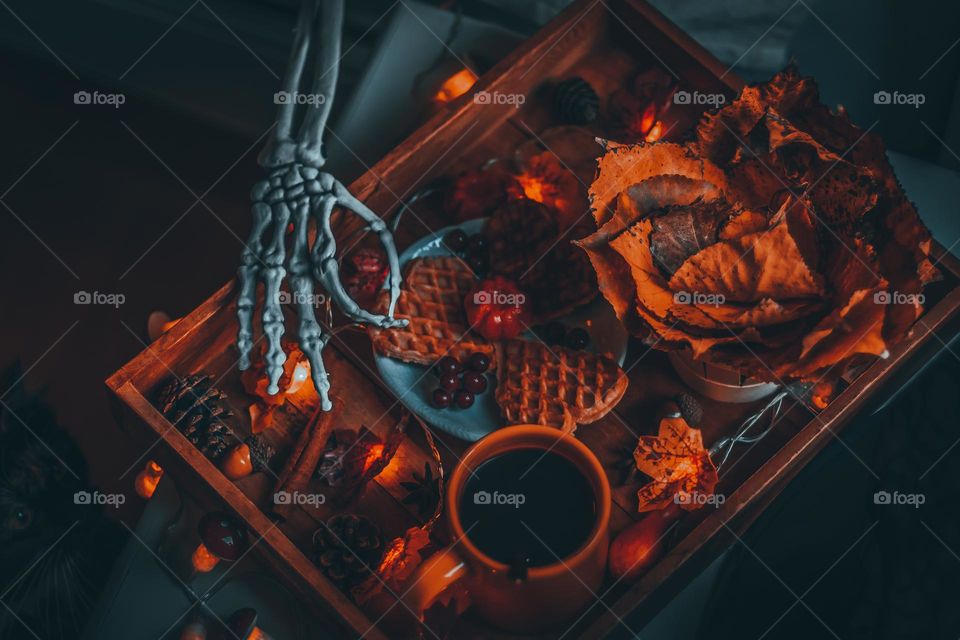 A skeleton hand stretching out from under the blanket on the bed at breakfast in a wooden tray standing on the bedside table with swafers, coffee, a bouquet of autumn leaves and a burning garland, close-up view from above. Halloween breakfast concept