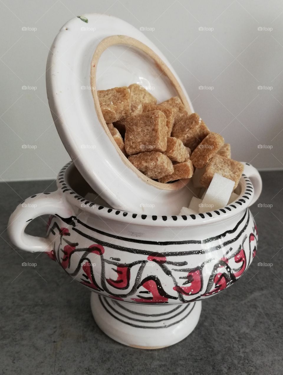 Multicoloured and patterned sugar container with handles and striped leg is full of pieces of white and brown sugar