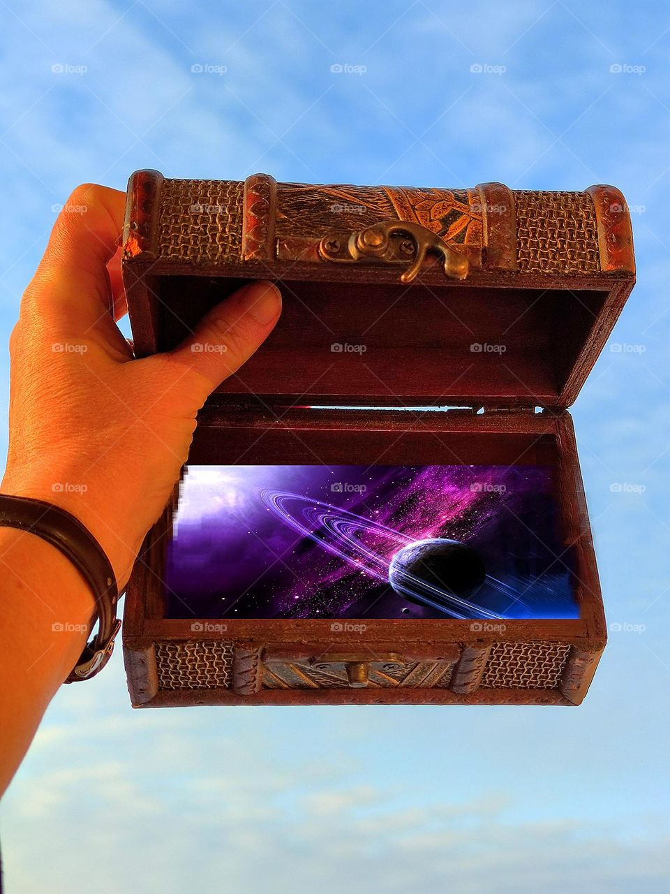 Surprise.  A hand holds a wooden antique box against a blue sky.  The lid of the box is open and the purple universe is visible.