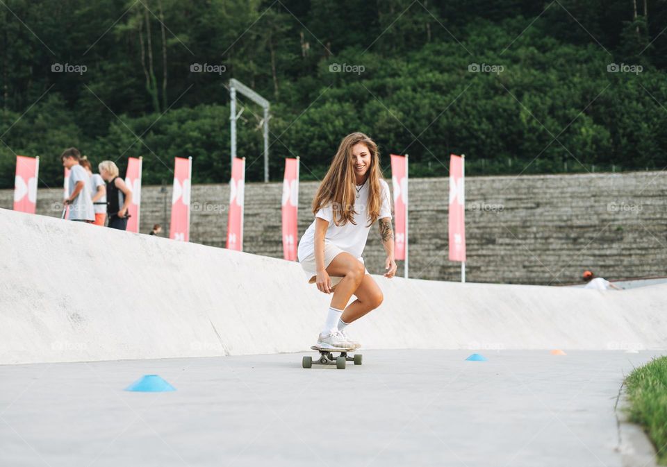 Slim young woman with long blonde hair in light sports clothes on longboard in outdoor skatepark at sunset time