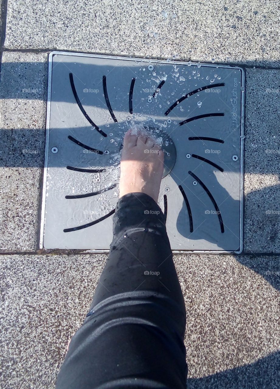pie de mujer mojado por chorro de agua de una fuente que se encuentra en el piso, en un parque