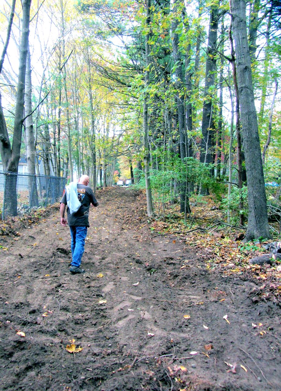 Man walking on dirt road in warm Autum day. His jacket is over shoulder. Overcast warm day hiking in woods. He's wearing street clothes, boots, t-shirt & long sleeve top over shoulder.